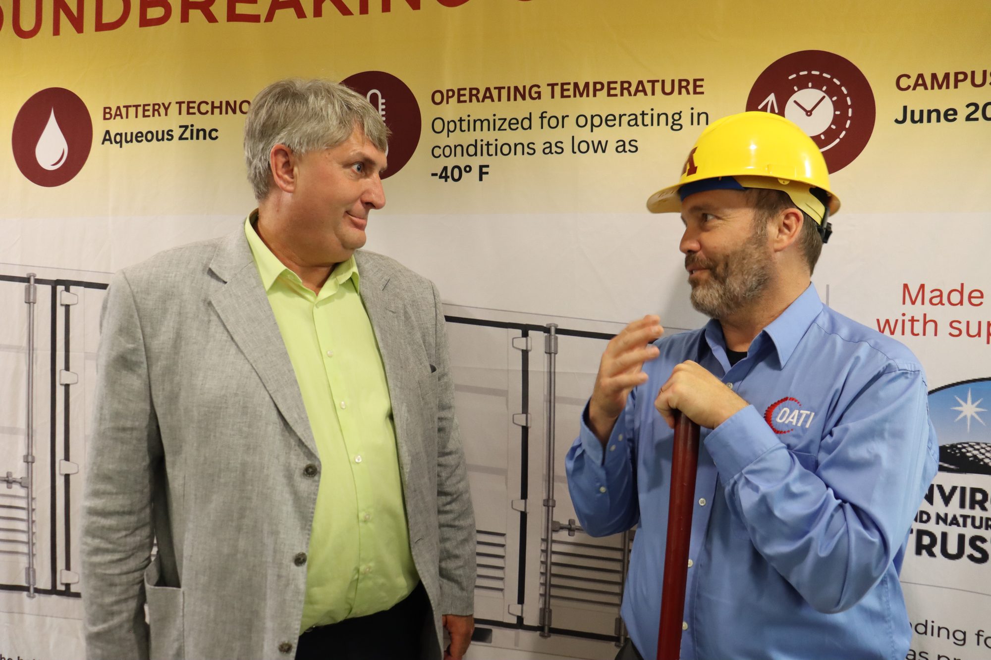 David Heim in a University of Minnesota hard hat talks with a guest in front of the banner